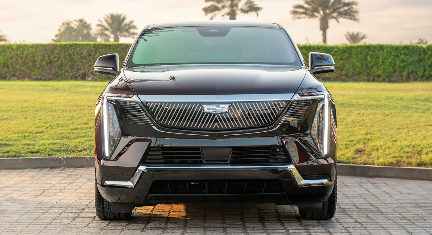 A sleek Cadillac Escalade IQL black SUV with a distinctive grille is parked on paved ground, surrounded by lush green grass and palm trees under a soft morning light.